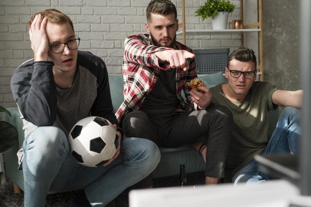 Como funciona o recurso de cashout em apostas esportivas? Confira! 3 3 homens sentados em sofá, segurando uma bola de futebol e apontando para frente com atenção.