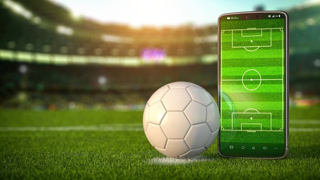 Como funcionam as apostas esportivas: é seguro? Vale a pena? 6 Uma bola de futebol ao lado de um celular com uma imagem de um campo de futebol, e um estádio ao fundo